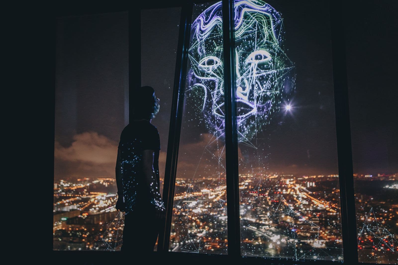A person beside a panoramic window at Cyber Zen, where a luminous digital portrait overlays the night-time city view.