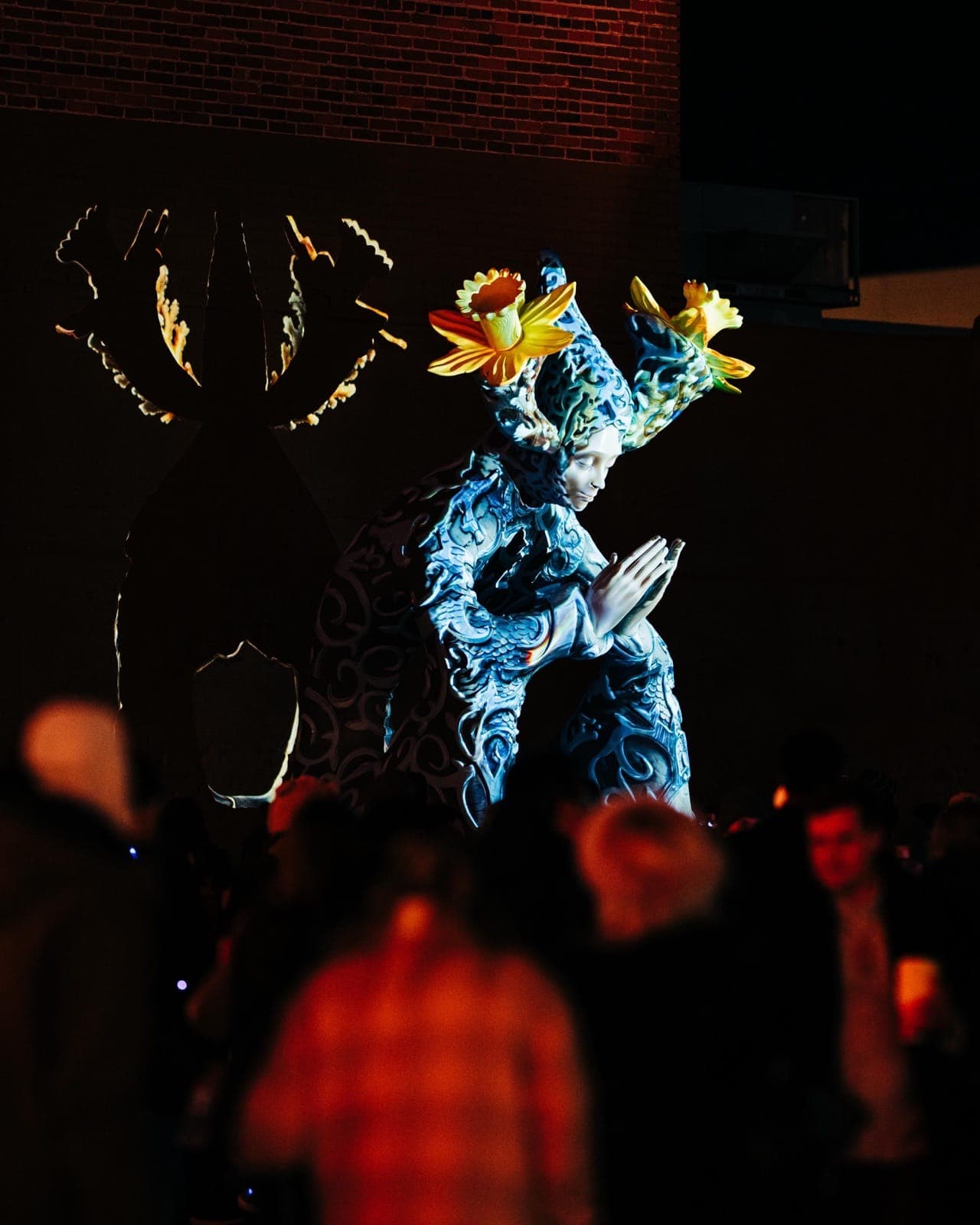 Daniel Popper's Ephemera at night — kneeling praying figure in cool blue baroque projection, golden flower crown lit warm, audience silhouettes in foreground, sculpture's shadow cast on the brick wall behind.