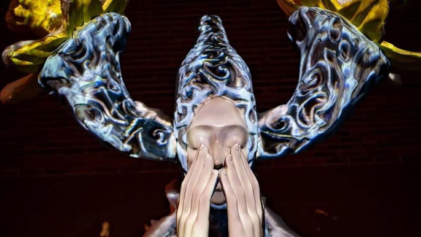 Final state — Ephemera lit at night, hands clasped at the face, baroque blue projection across the robe, golden flowers on the antler crown. Photo by Tyreek B Hitt.