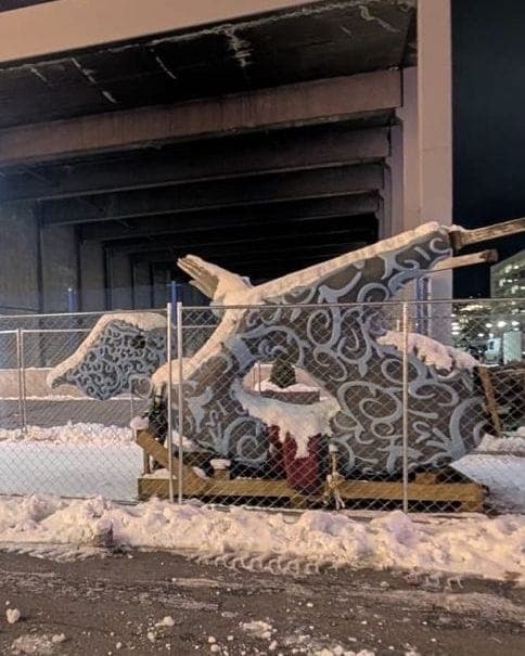 Setup night — the sculpture's base mid-rig, surrounded by piles of snow, a forklift visible in the foreground, the parking-garage volume of downtown Reno behind.