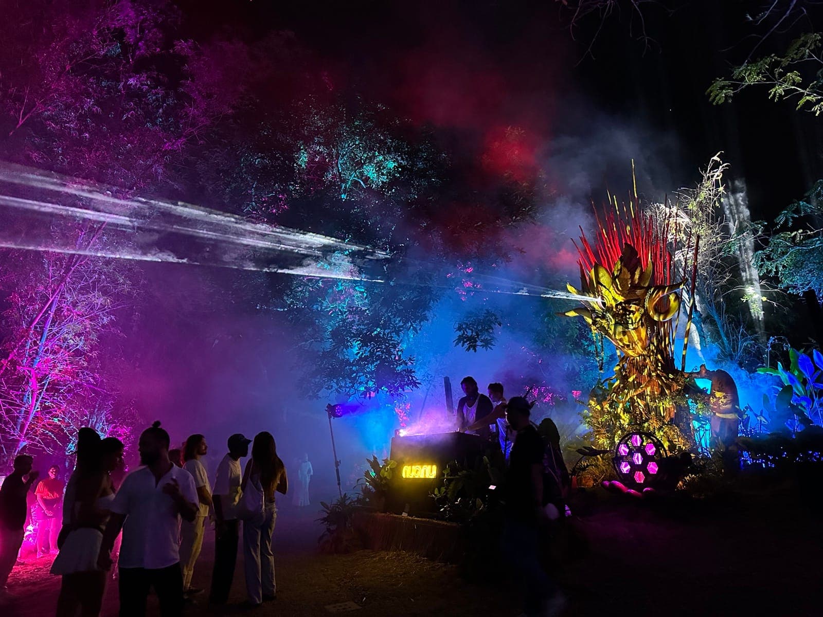 Daniel Popper's Kriya at night, lit purple and yellow with smoke and laser beams across the jungle, audience gathered at the base.