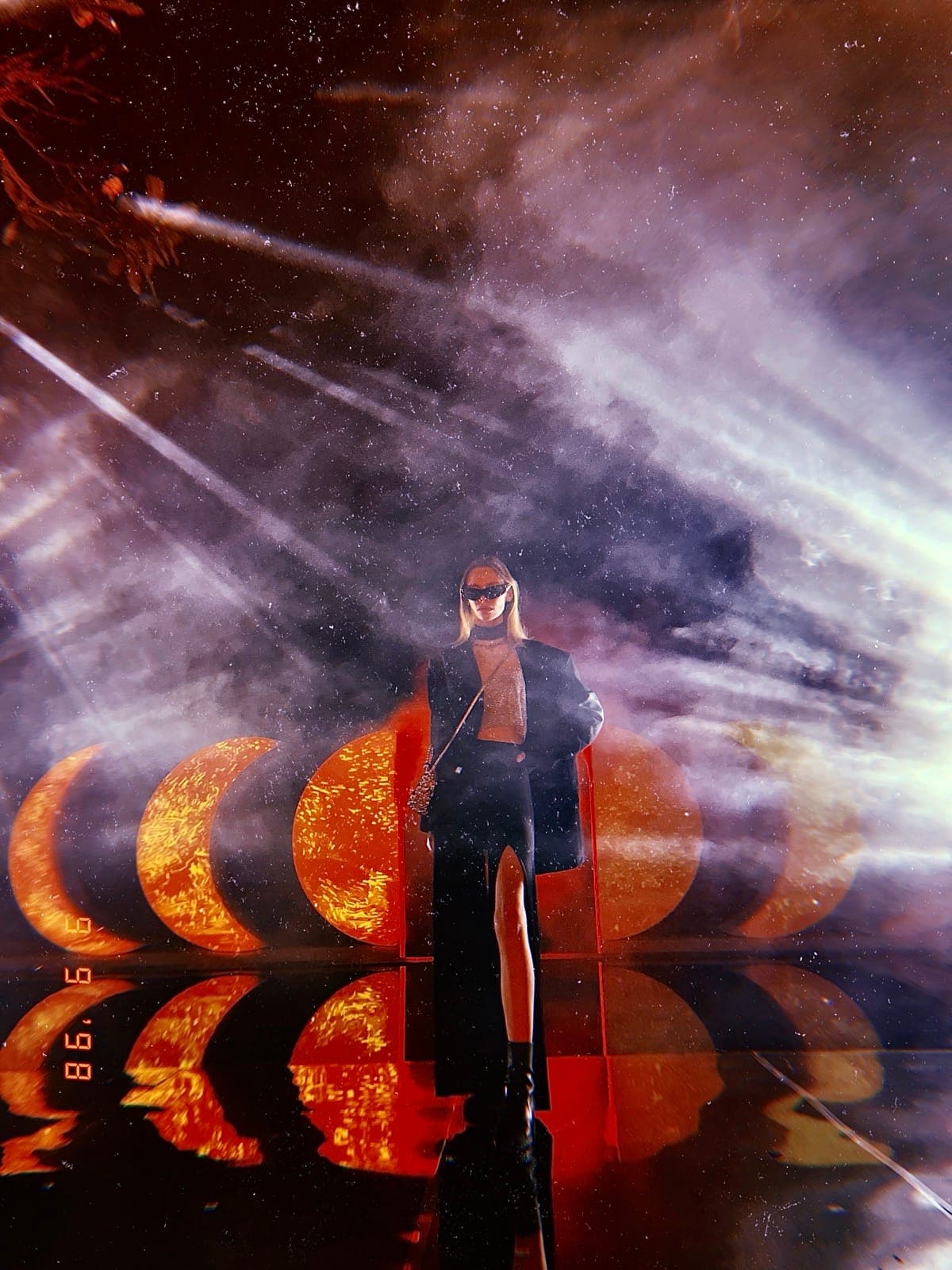 Moon Phases at night — a figure in sunglasses framed by laser smoke, the five crescent moons lit warm orange behind, mirrored on the wet floor.