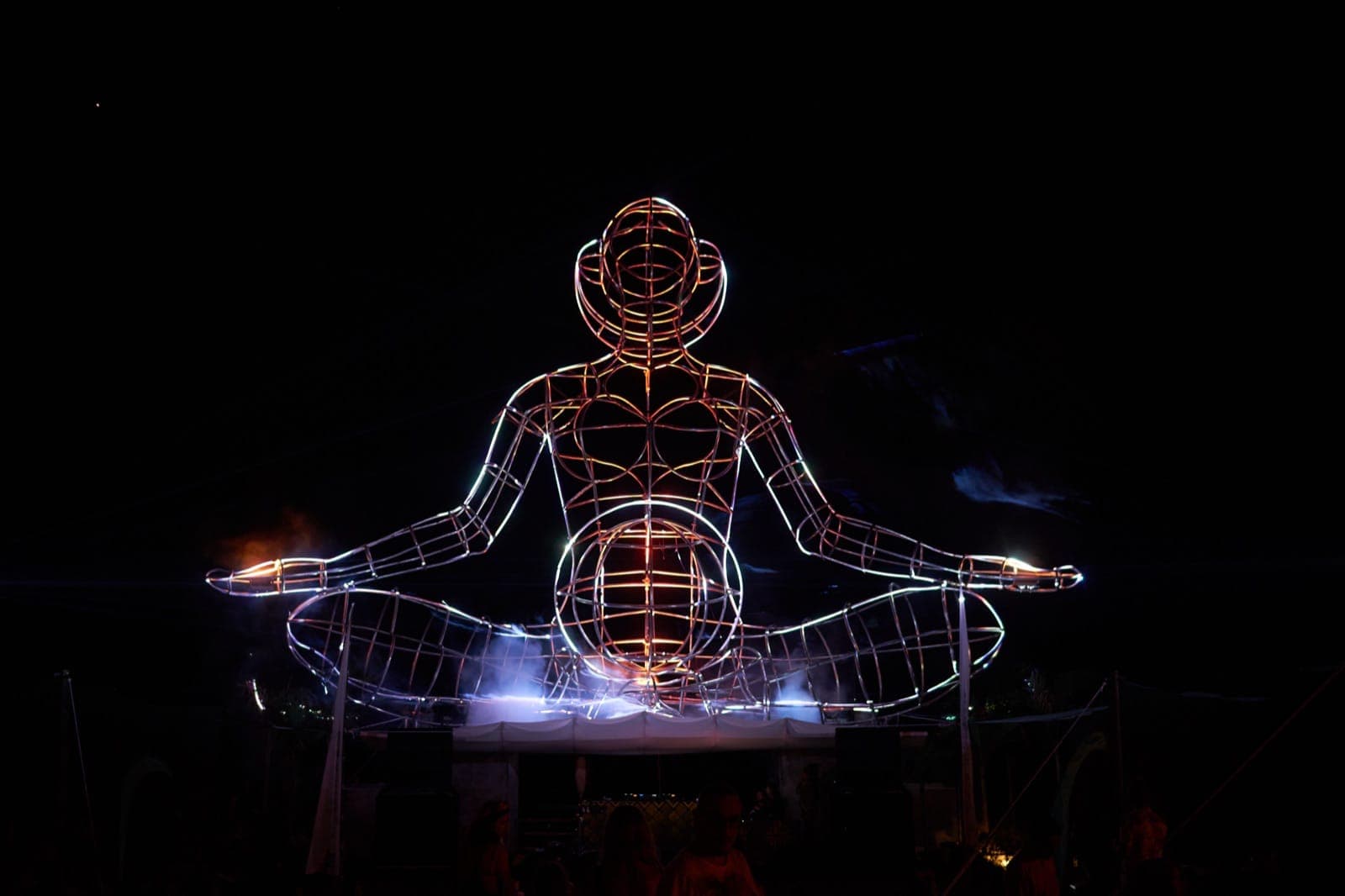LUNA sculpture lit pink and red at night, seated in lotus, audience below.