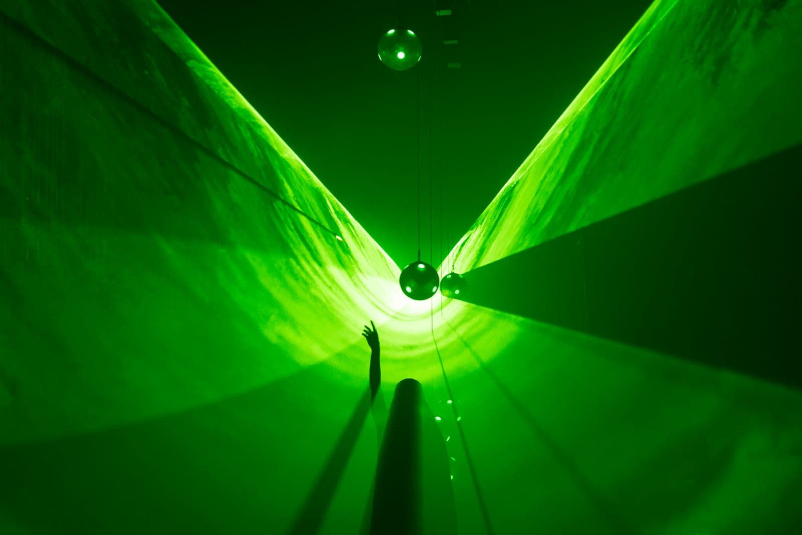 A visitor reaches into the green projection field beneath suspended mirrored spheres